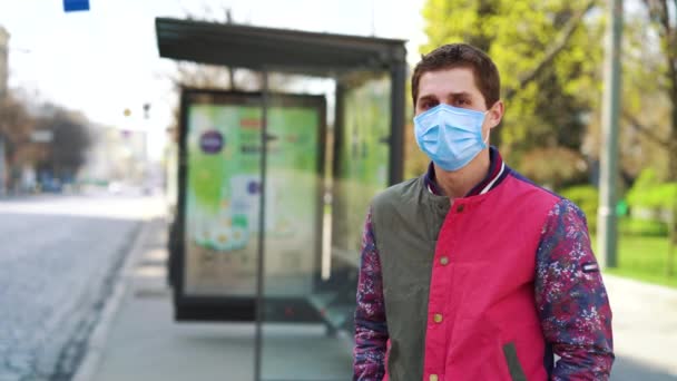 Junger Mann mit medizinischer Maske wartet an leerer Bushaltestelle auf Bus — Stockvideo