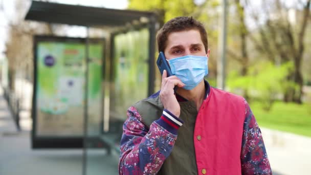 Mladý muž v lékařské masce mluví po telefonu na autobusové zastávce — Stock video