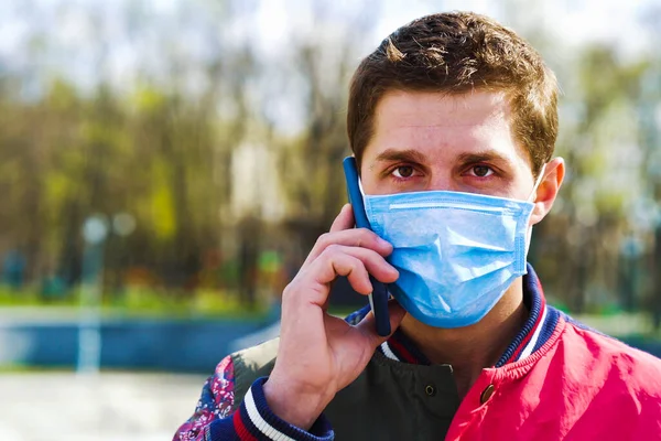 Tıp maskeli genç adam parkta cep telefonuyla konuşuyor.