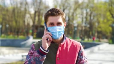 Koruyucu maskeli genç adam parkta yürüyor ve telefonla konuşuyor.