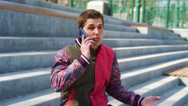 Good-looking young man talking on mobile phone on concrete stairs outside — Stock Video