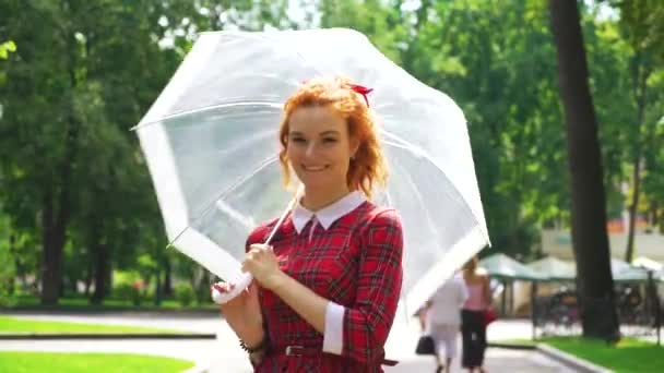 Positive red-haired girl walking in green park with umbrella on sunny day — Stock Video