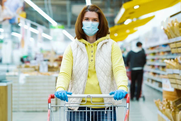 Maskeli ve eldivenli genç bir kadın süpermarkette alışveriş arabasıyla