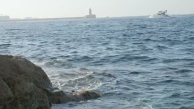 Yelkenli tekne sağda mavi su yaklaşıyor. Bazı son dakika dalgalar ile kayalık sahil ön plan vardır.