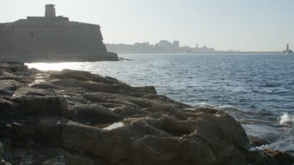 Sailing boat approaching from right on blue water. Foreground has rocky shore with some breaking waves. — Stock Video