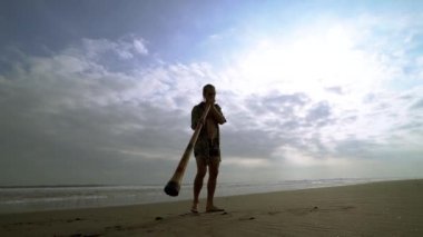 Genç müzisyen didgeridoo ve gitarlı kız gün batımında plajda çalıyor..