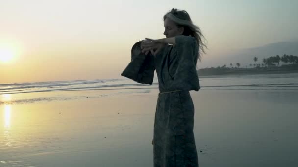 Blanke vrouw gekleed in een kimono dansen door de oceaan bij zonsondergang — Stockvideo