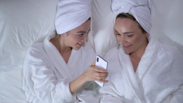 Chicas tumbadas en la cama y el uso de la aplicación móvil de compras en el teléfono inteligente, teniendo fiesta de spa — Vídeos de Stock