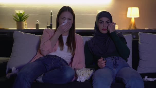 Két lány szomorú filmet néz a kanapén, sírnak és könnyeket törlik, együtt szórakoznak.