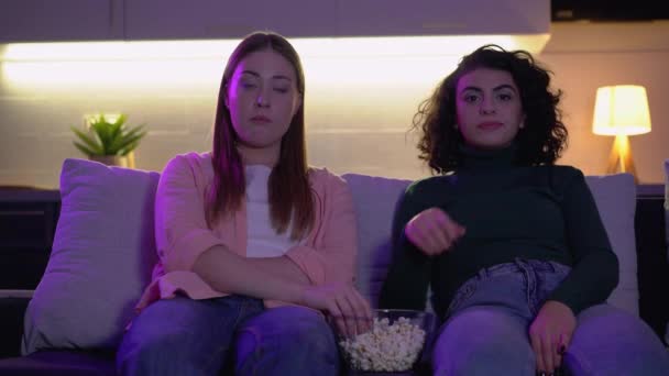 Две женщины жуют попкорн, смотрят телевизор на диване дома, ленивый образ жизни — стоковое видео