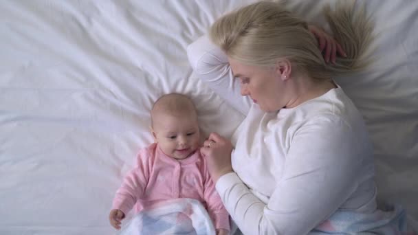 Mutlu anne yatakta bebekle yatar aktif bebek battaniyeyle oynar, şekerleme yapar — Stok video