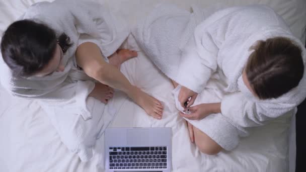 Chicas adolescentes sentadas en la cama, viendo películas en línea, pintando uñas de los pies, cuidado de la belleza — Vídeos de Stock