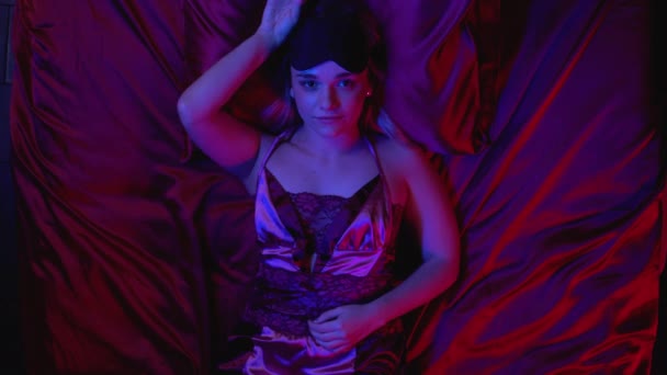 Γοητευτική νεαρή κοπέλα που φοράει μεταξωτή πιτζάμα ξαπλωμένη στο κρεβάτι μόνη, κοιτάζοντας την κάμερα — Αρχείο Βίντεο