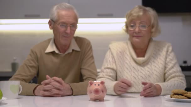 Oudere echtpaar zetten munt in piggybank, op zoek naar camera, planning budget — Stockvideo