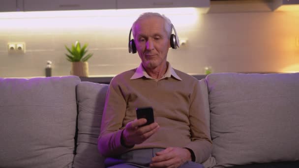Старший слушает музыку в беспроводных наушниках, держа мобильный, наслаждаясь — стоковое видео