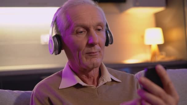 Пожилой европеец наслаждается любимой музыкой, носит беспроводные наушники, гаджет — стоковое видео