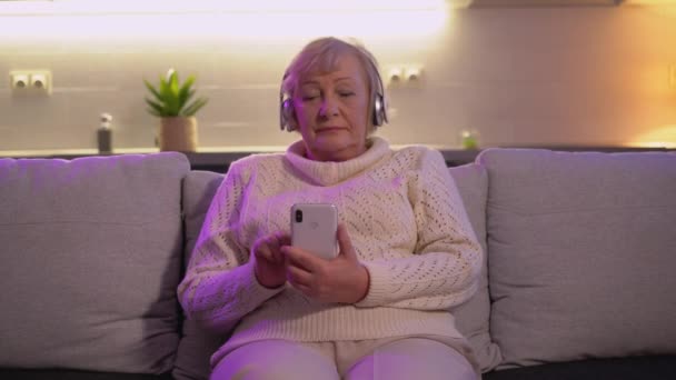 Positive elderly woman in wireless headphones enjoying music, using phone app — Stock Video