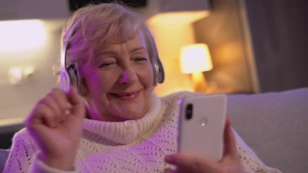 Ευτυχισμένη ηλικιωμένη γυναίκα που παρακολουθεί μουσικά βίντεο στο τηλέφωνο, χρησιμοποιώντας ασύρματα ακουστικά — Αρχείο Βίντεο