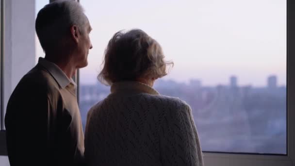 Äldre gifta par står vid fönstret tillsammans, omtänksam make kramar fru — Stockvideo
