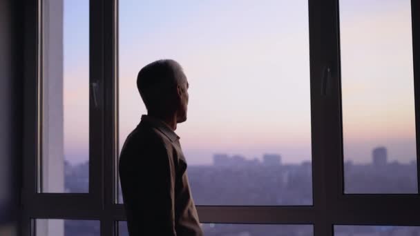 Δυστυχισμένος ηλικιωμένος άντρας κοιτάζει στο παράθυρο, νιώθει μοναξιά στην πόλη, λυπημένες αναμνήσεις — Αρχείο Βίντεο
