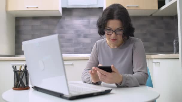 Orta yaşlı bir kadın evde akıllı telefon ve iş başvurusu üzerine makale okuyor. — Stok video