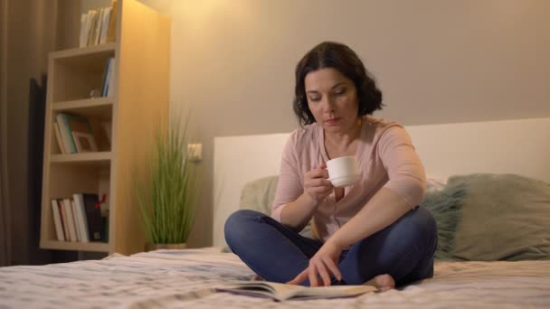 Mujer de mediana edad leyendo libro y tomando té, relajarse en la cama en casa, descansar — Vídeos de Stock