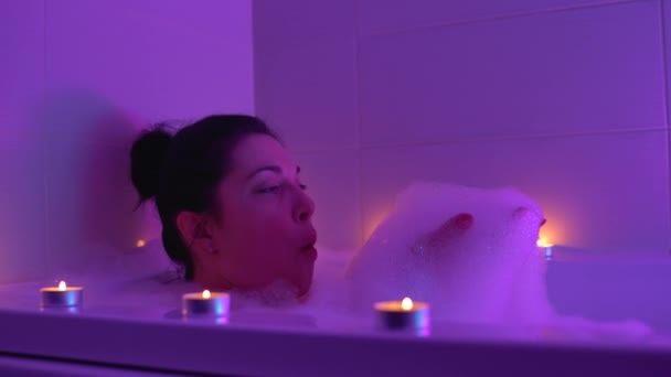 Hravá šťastná žena fouká do pěny, leží ve vaně se svíčkami, lázeňský relax — Stock video