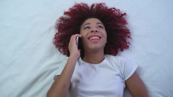 Счастливая африканская женщина разговаривает по телефону, лежит в постели дома, общение — стоковое видео