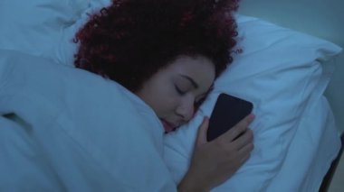 Afrika kökenli Amerikalı genç yatakta uyuyor, elinde akıllı telefon, alet bağımlılığı var.