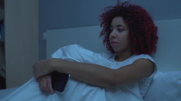 Triste adolescente avec smartphone à la main assise sur le lit la nuit, adolescence — Video