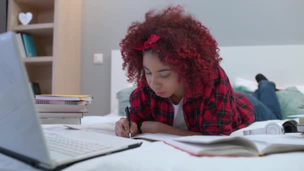 Estudiante de raza mixta escribiendo en cuaderno, haciendo deberes, acostado en la cama en casa — Vídeos de Stock