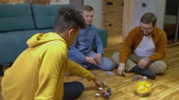 Молодой человек, раздающий карты, друзья, играющие в покер дома, сидящие на полу — стоковое видео