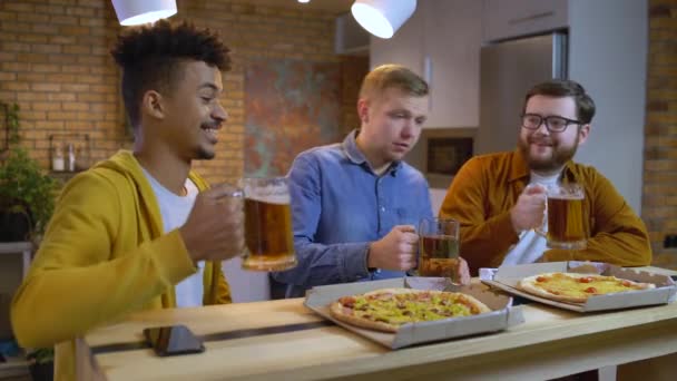 Nadšení muži slaví, cinkají sklenicemi piva doma, pizza na stole — Stock video