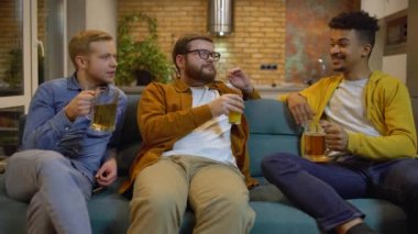 Çok kültürlü erkek arkadaşlar kadehleri içeceklerle tokuşturur, kanepede sohbet ederler.