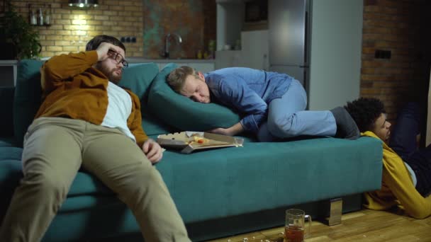 Пьяный молодой человек просыпается после вечеринки, друзья спят в грязной комнате, похмелье — стоковое видео