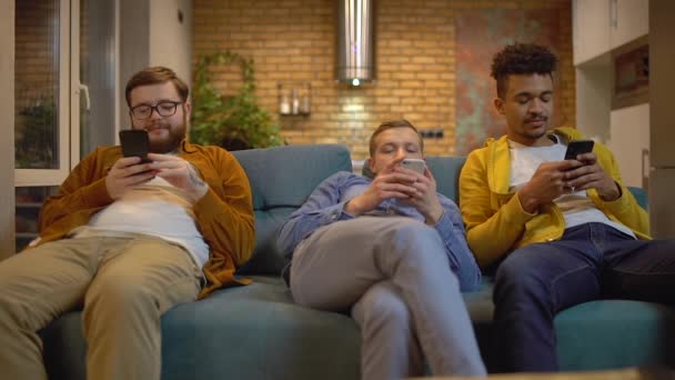 Tres hombres jóvenes navegando sitios en teléfonos inteligentes, sentado en el sofá, adicción gadget — Vídeo de stock