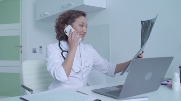 Médica feliz chamando paciente por telefone, olhando para a ressonância magnética, boas notícias — Vídeo de Stock