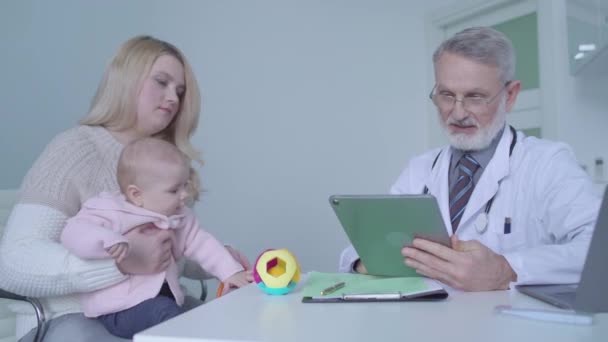 Семейный врач консультирует молодую мать с ребенком ведения медицинских записей в планшете — стоковое видео