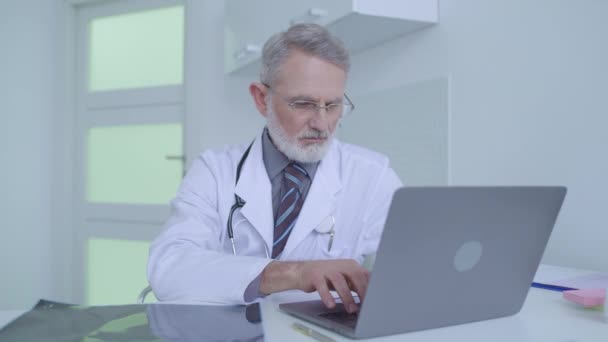Médico senior revisando la resonancia magnética cerebral, escribiendo diagnóstico en el portátil, medicina — Vídeo de stock