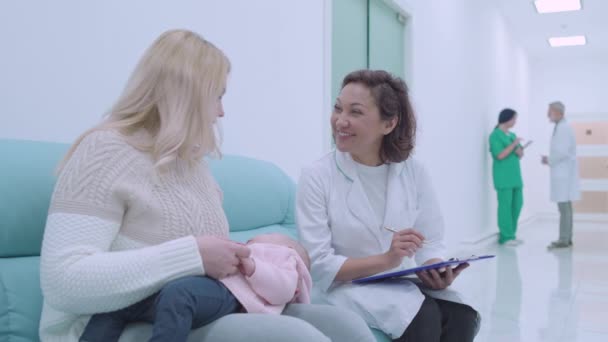 Дружня жінка-лікар заповнює документи, розмовляє з пацієнткою з дитиною — стокове відео