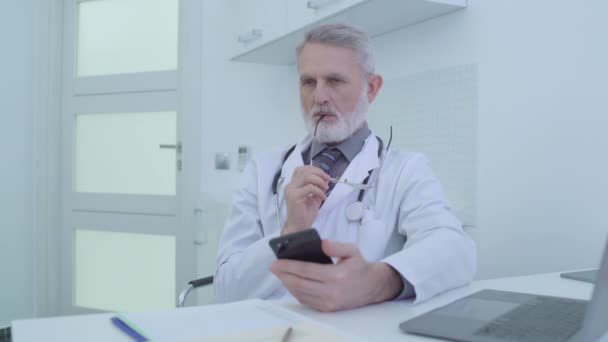 Pensionär läkare på kontoret med smartphone, funderar över behandling för patienten — Stockvideo