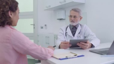 Doktor tableti tutuyor, klinikte kadın hastayla konuşuyor, teşhisi tartışıyorlar.