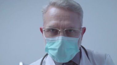 Gözlüklü ve koruyucu maskeli bir erkek doktor kameraya bakıyor.