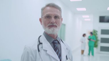 Laboratuvar önlüklü gri saçlı doktor kameraya, tıbbi servise, hastaneye bakıyor.