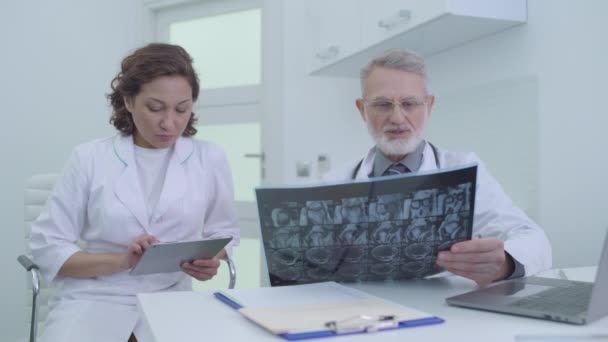 Врач осматривает пациентов рентгеновским снимком, медсестра заполняет медицинские карты на таблетках — стоковое видео