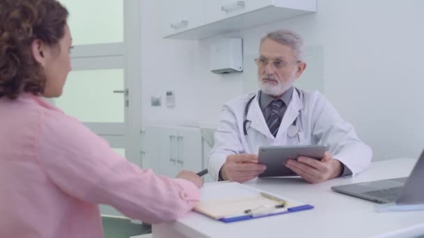 Doktor tableti tutuyor, klinikte kadın hastayla konuşuyor, teşhisi tartışıyorlar. — Stok video
