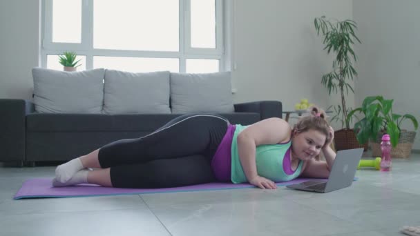 Lui overgewicht meisje surfen internet op laptop, liggend op mat in plaats van uit te oefenen — Stockvideo