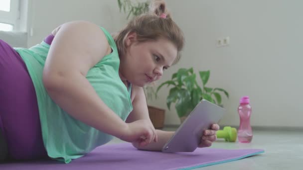 Plus storlek ung kvinna rulla tablett, ligger på mattan, söka fitness tips — Stockvideo