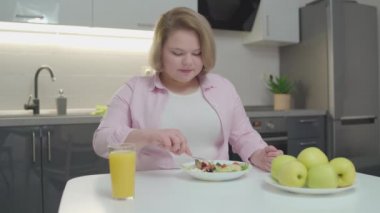 Tombul kadın mutfakta taze sebze salatası, masada elma ve meyve suyu yiyor.
