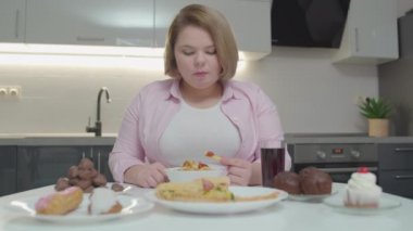 Obez kadın patates kızartması yiyor ve masada soda, pizza ve kek içiyor.
